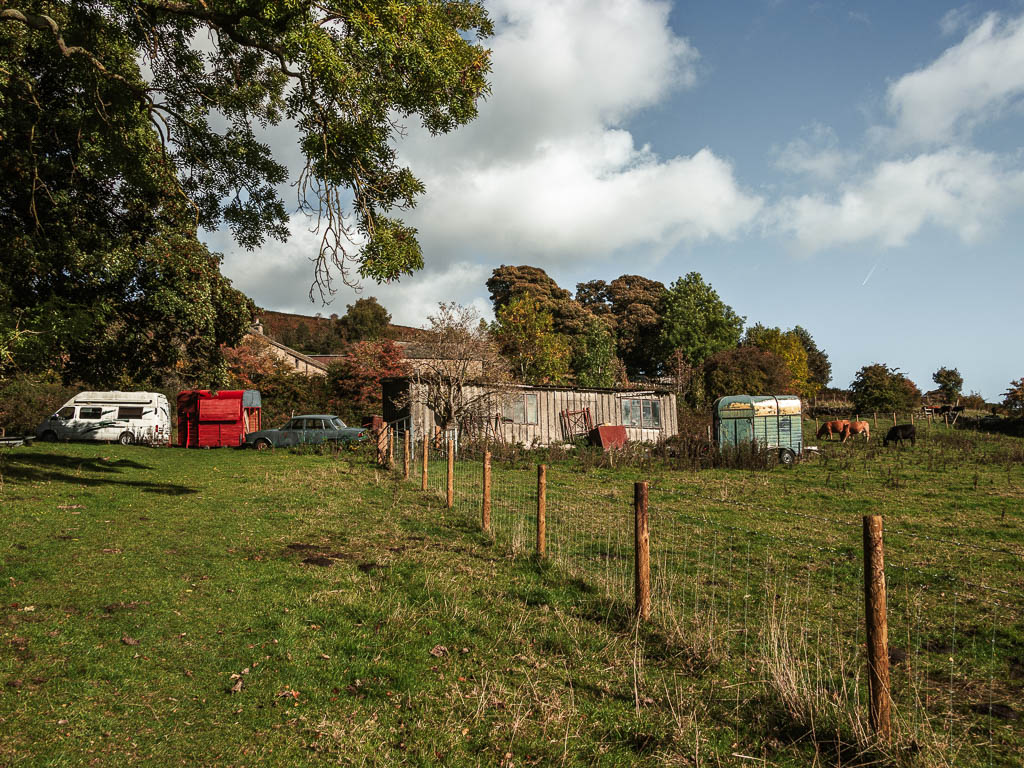 Looking up the green grass hill, with a metal fence eon the left, towards metal sheds and some parked cars ahead.