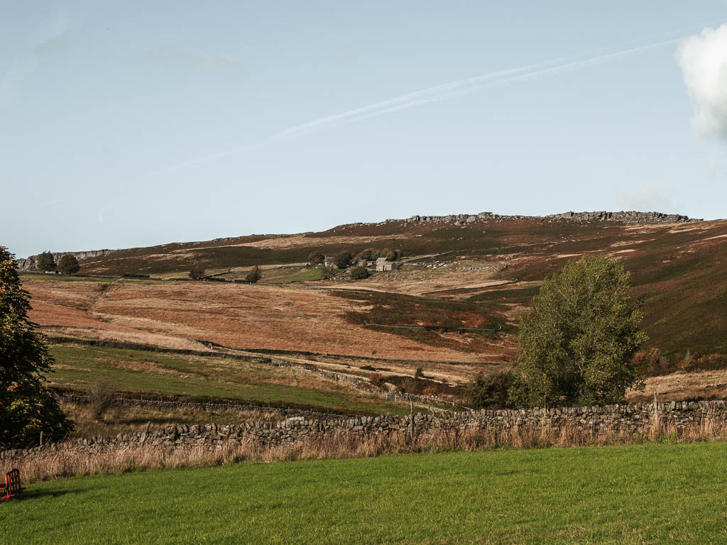 Looking across the grass field to a stone wall, and a big hill beyond with a view to both Higger Tor and Stanage Edge on top of the hill, near the end of the circular walk route.