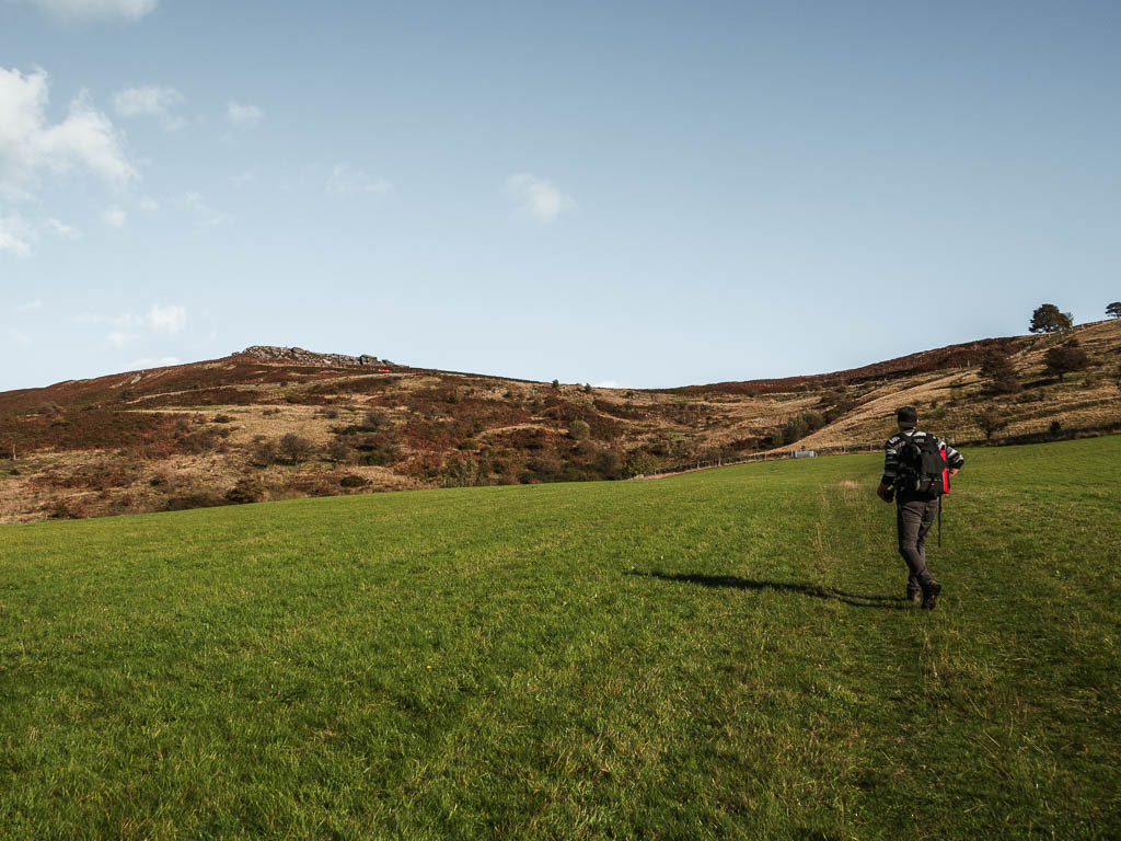 A large grass field hill, with a view to higger tor ahead on the hill, near the end of the circular Stanage Edge walk. There is a man walking up the hill.