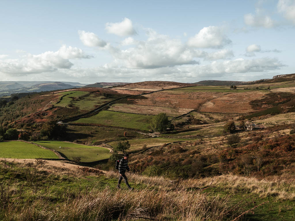 Looking down to the valley and undulating hills near the end of the Stanage Edge circular walk. There is a man walking across the hill.