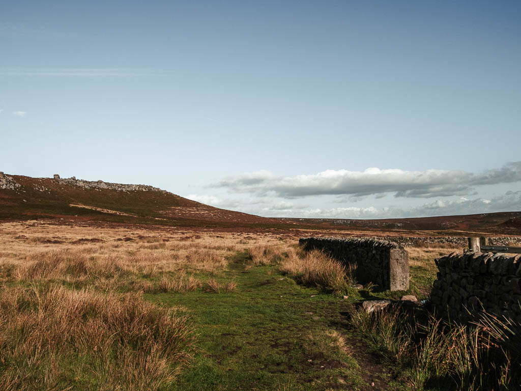 A grass path with a stone wall on the right, and Higger  Tor rising up ahead to the left.