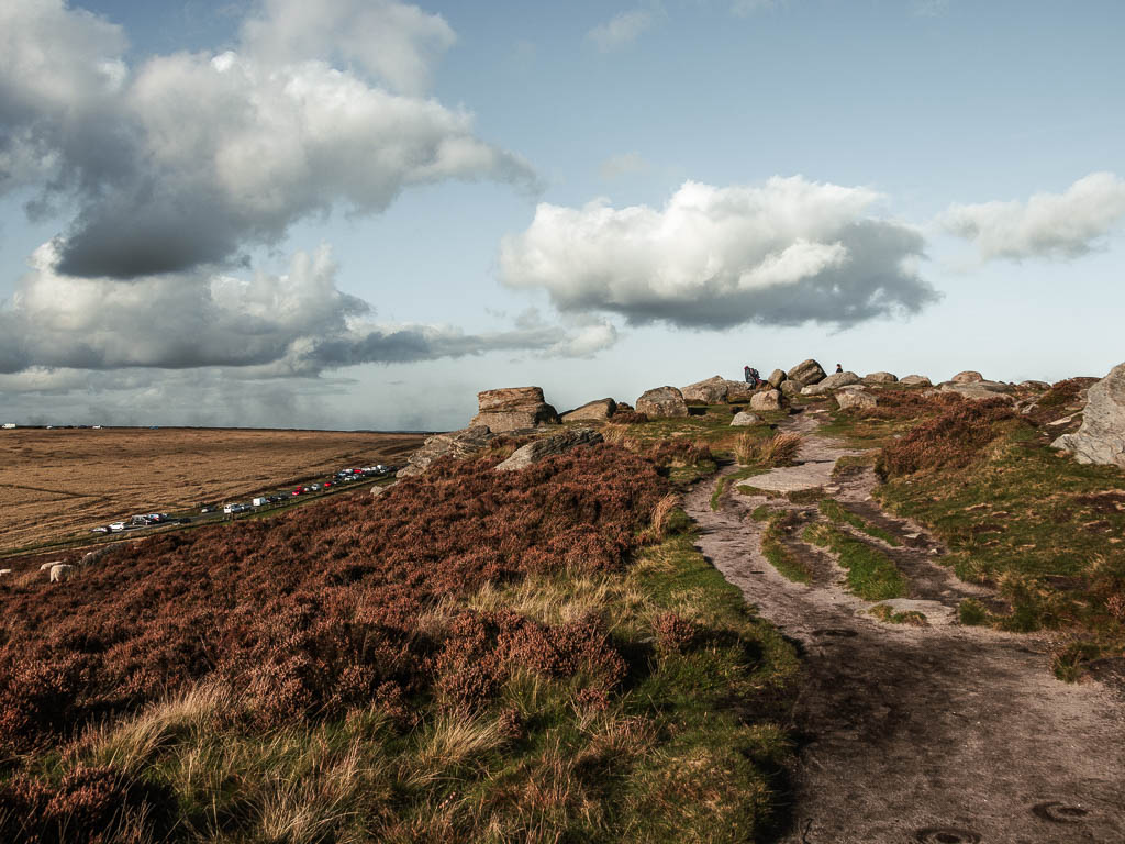 A dirt path leading to large rocks, with a view down the hill to the left of the road lined with parked cars.