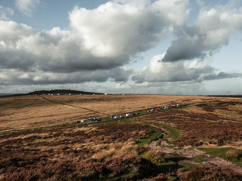 Looking down the hill, to a road lined with parked cars, and a large field on the other side with a trail running through it. near the end of the Stanage Edge circular walk.