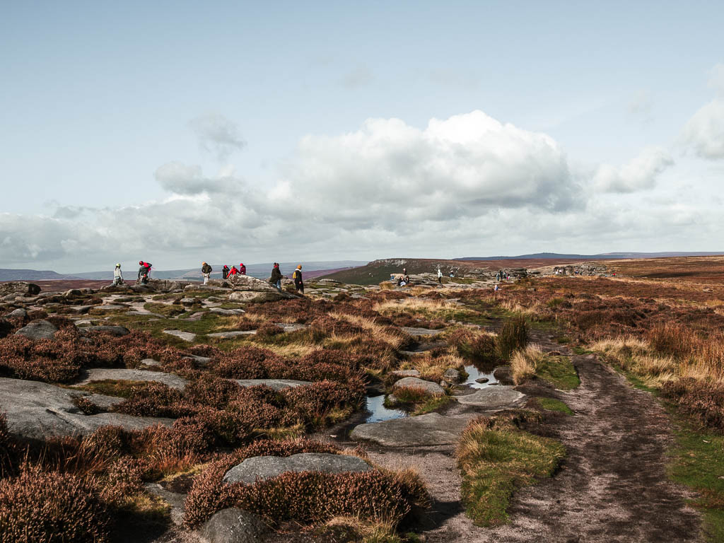 Walking along the top of Stanage Edge with flat rocks and tufts off grass in between. There is a group of people ahead.