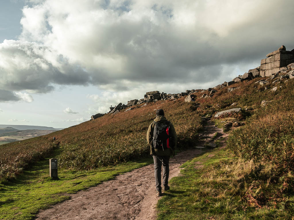 A man walking along a dirt trail, leading up to the rocks of Stanage Edge. There is a small grass trail leading off the dirt trail to the left.