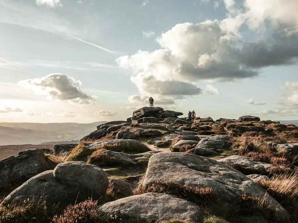 A mass of boulders rising up ahead, with a person standing on top of the highest one in the Peak District National Park.