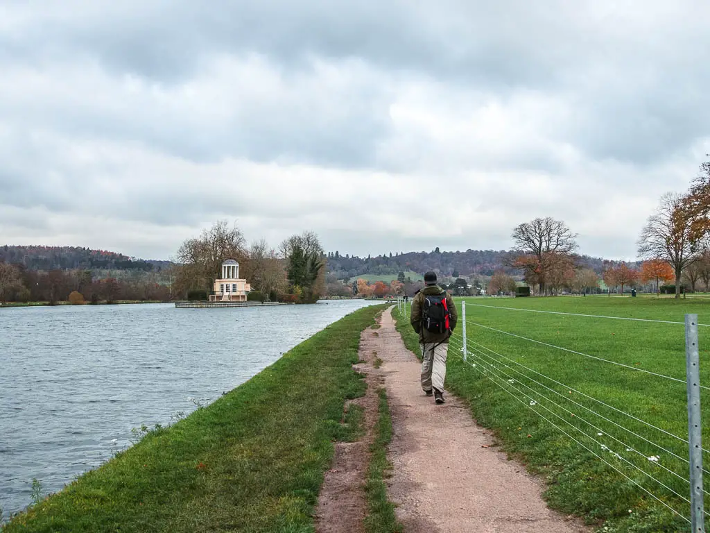 A man walking along a path, with a grass bank to the left then the river thames, on the walk from Henley to Hambleden. There is a wire fence to the right, temple island ahead, and the hills beyond that.