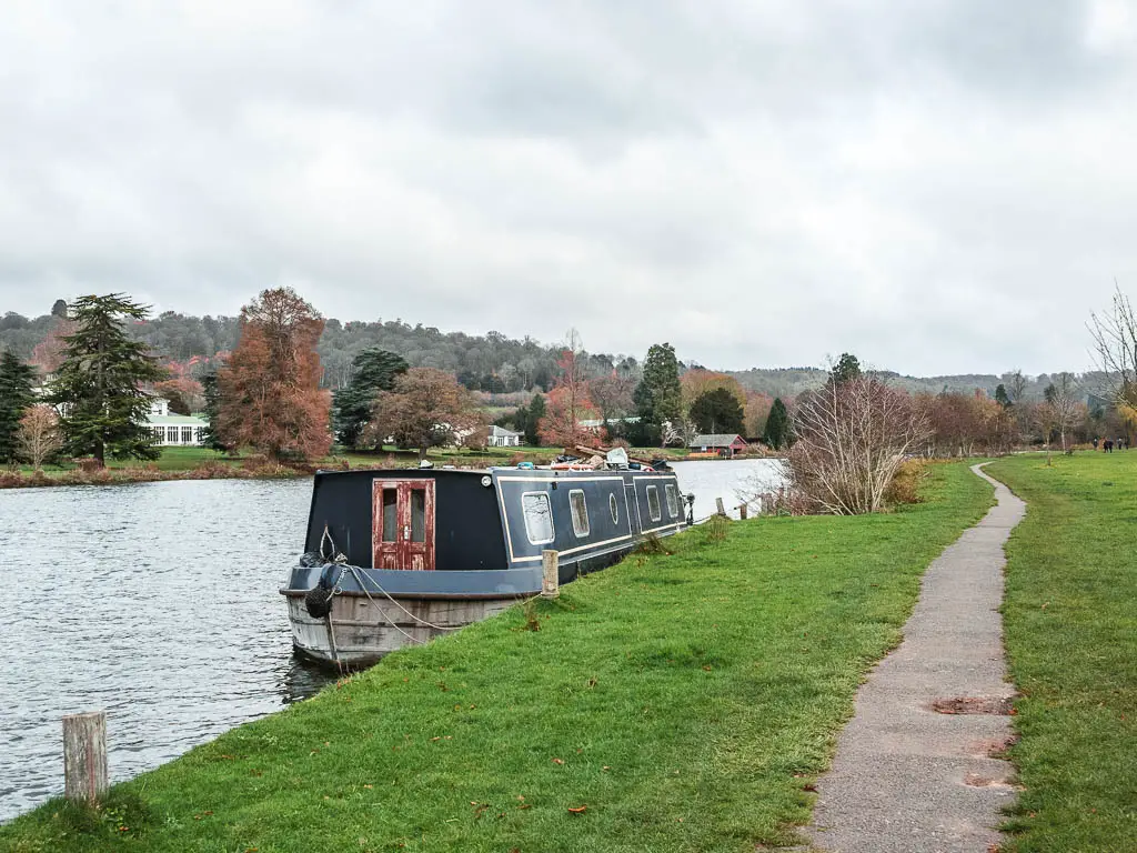 A path along the side of the river thames, with a barge moored to the side, on the walk from Henley to Hambleden.
