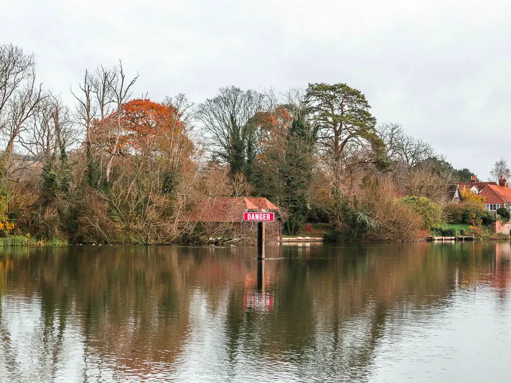 A red danger sign in the middle of the river, with lots of leafless trees on the other side.