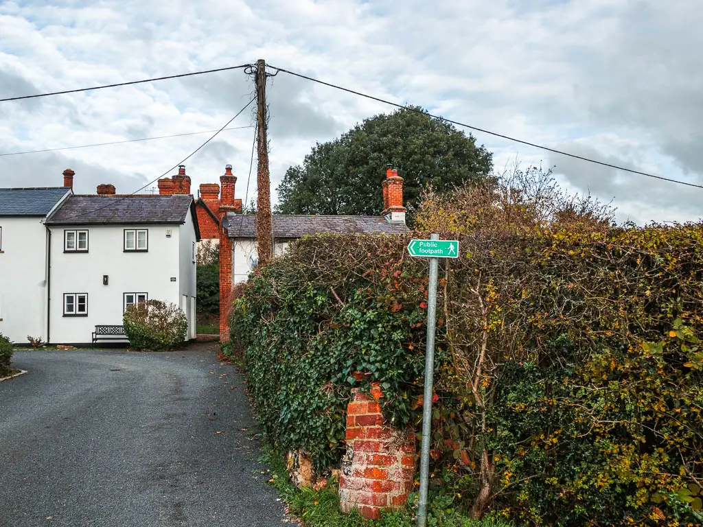 A trail signpost in front of a hedge, pointing left towards a group of houses with a gap between them.