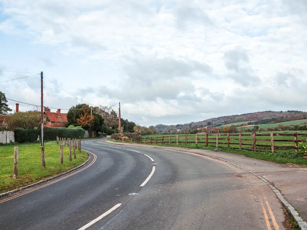 A road curving ahead, with a grass bank to the left and a green grass field to the right with a wooden fence.