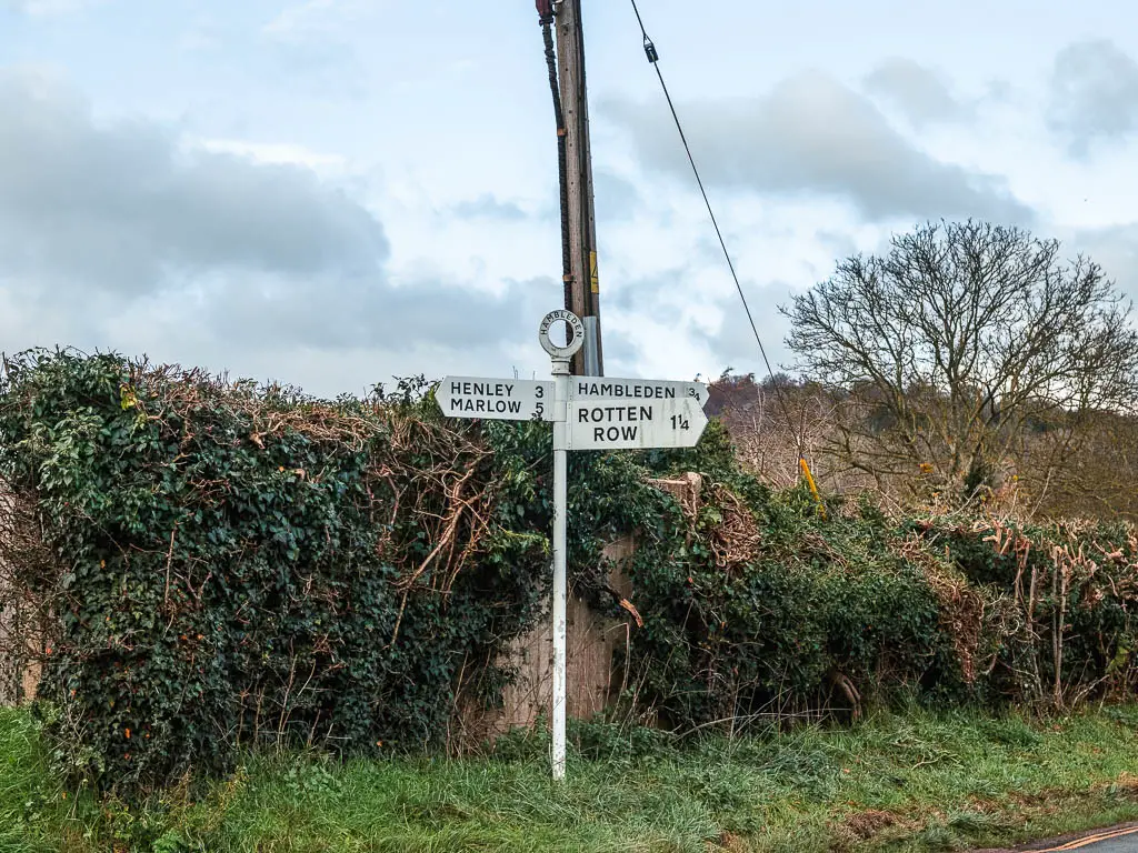 A white signpost in front of a hedge and wall. The signpost points to Rotten Row, Henley, and Marlow.