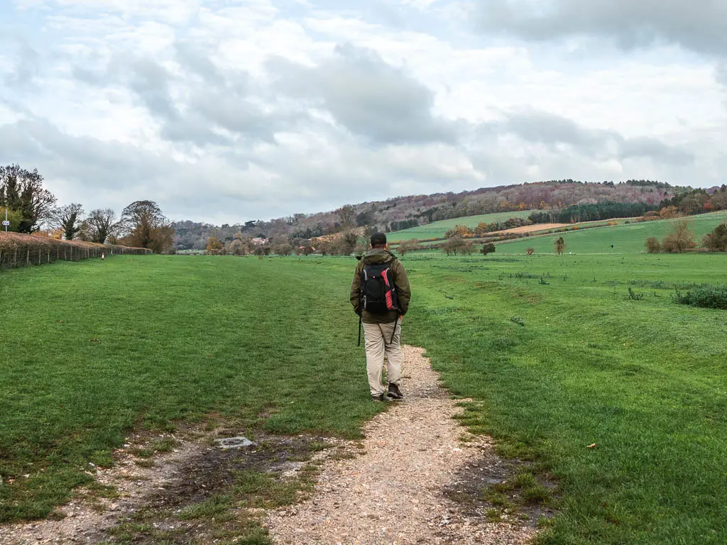 A man walking along a trail in the large field with neatly cut grass and hills ahead in the distance.