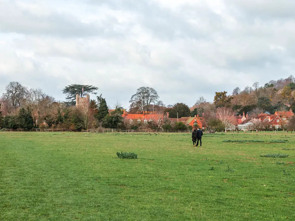 A large neatly cut grass field, with the cottages of Hambleden ahead, half way through the walk from Henley. There are a couple of people walking in the field ahead.