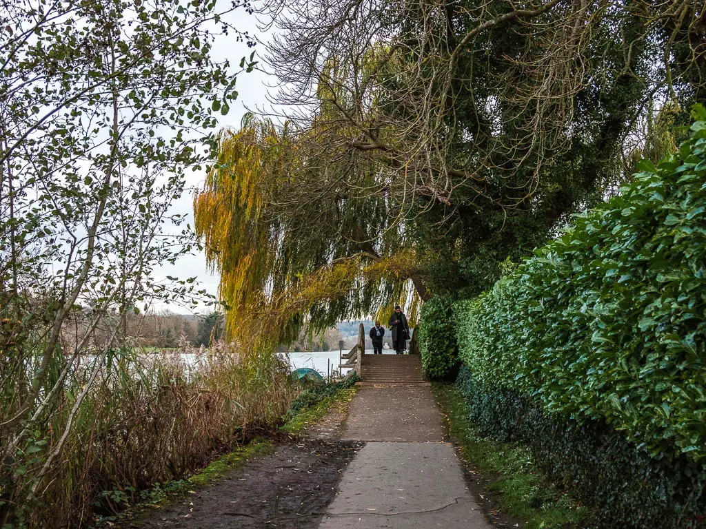 A path with a hedge to the right with over hanging trees, the river to the left, and a small bridge ahead with people walking over it.