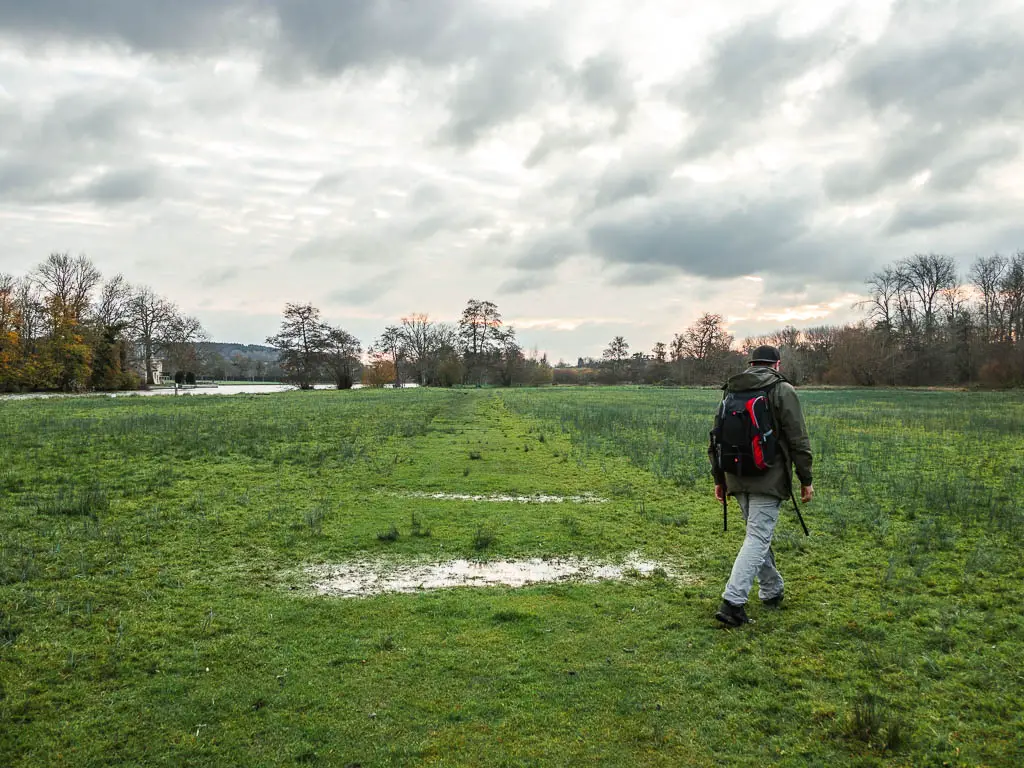 A large grass field, with a rough trail straight through it filled with puddles. A man is walking to the right of one of the puddles.
