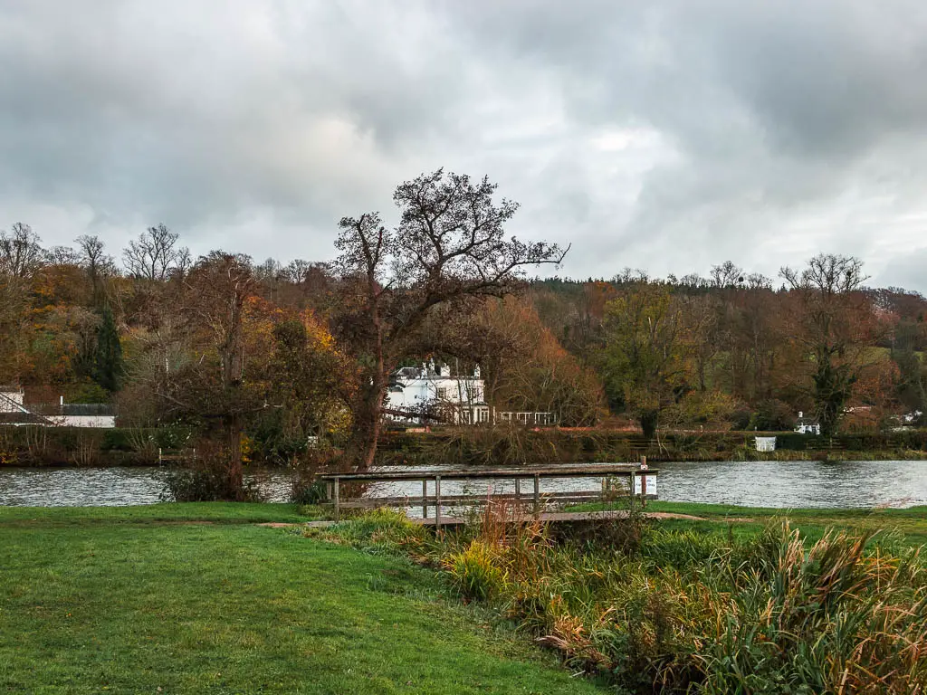 Looking towards a wooden bridge connecting two field, with the river thames on the other side and a white coloured house on the other side of the river.