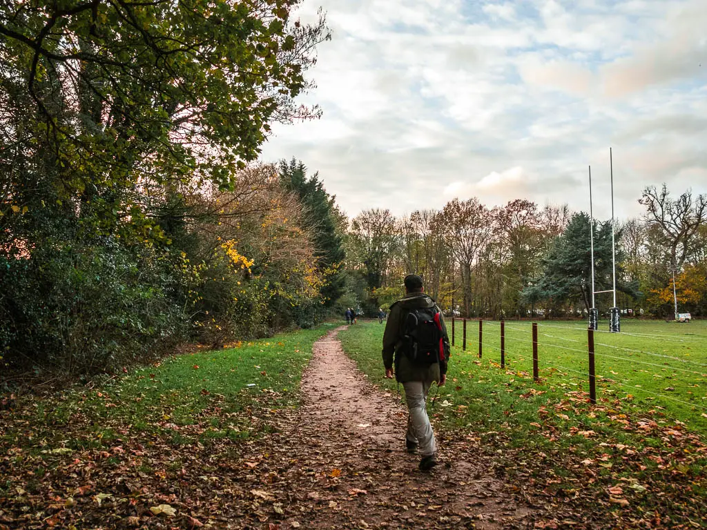A man walking along a trail through the grass, with trews and bushes to the left and a rugby field to the right.