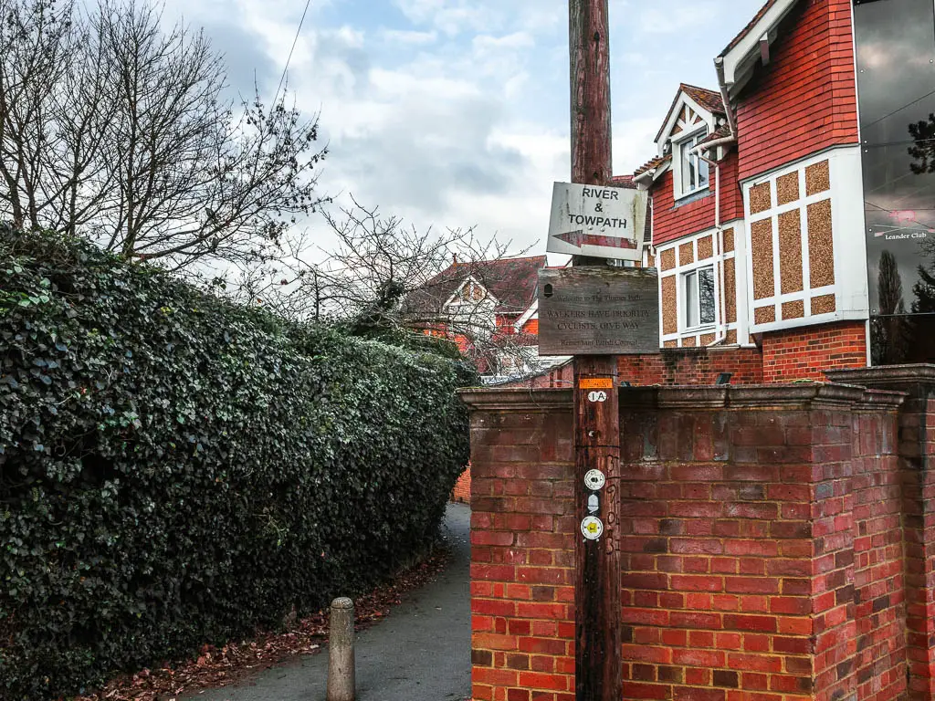 A path with a green hedge to the left, and brick wall and houses to the right. There is a sign on a post saying 'river and towpath' with a red arrow pointing along the path.