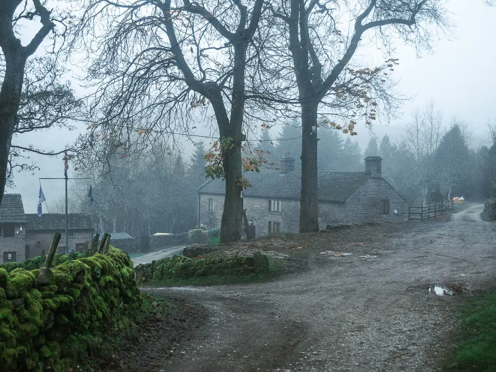 The road, with a moss covered stone wall on the left, and some buildings ahead. Its a dark very foggy day.