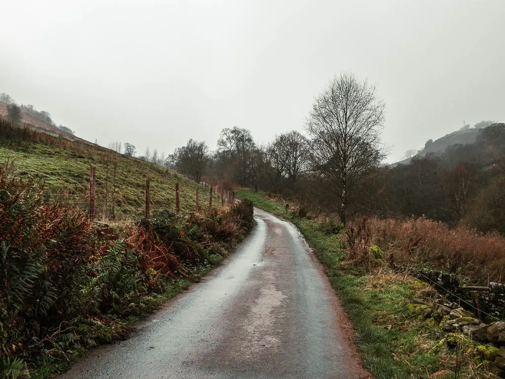 A road leading straight and curving to the left ahead, with a green grass hill and fence on the left.