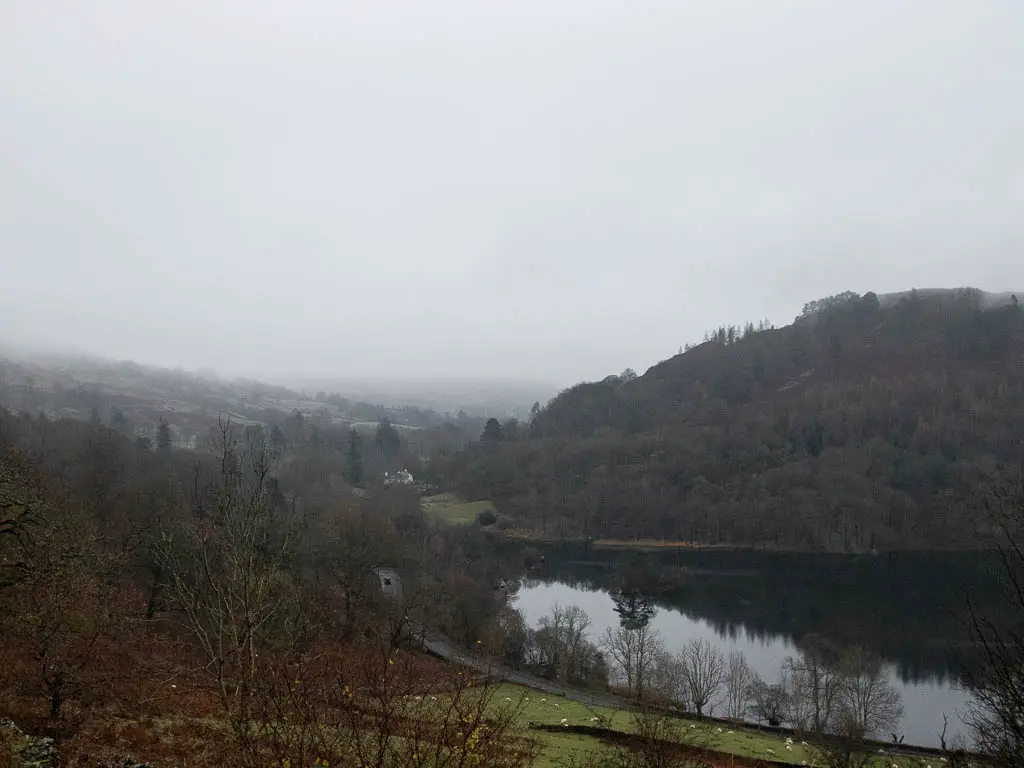 Looking down to Rydal Water on a foggy day, on the walk around it. There is a big tree covered hill on the other side. 