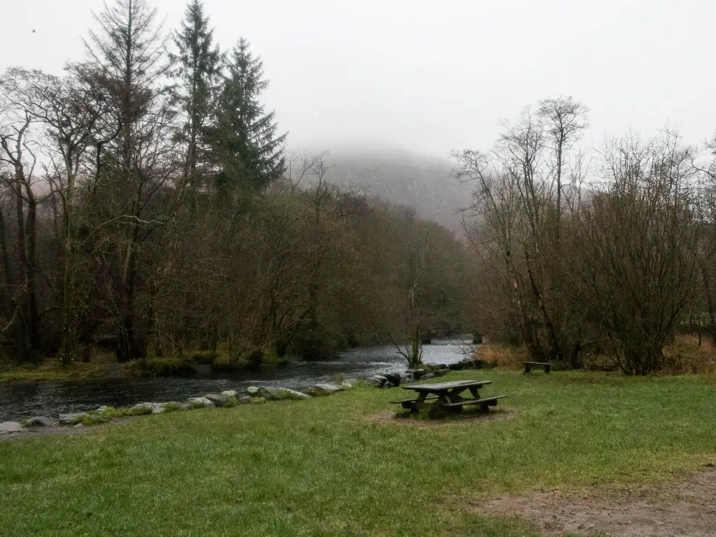 Looking across the grass towards the river on the Rydal water and cave circular walk. There is a picnic bench on the grass, and woodland trees on the other side of the river. 