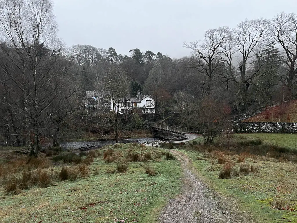 A gravel trail through the grass leading to a bridge, and a white building on the other side, on the Rydal water circular walk.