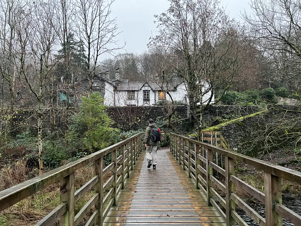 A man walking along the wooden bridge with a stone wall ahead, and a white budding behind it, on the way to Rydal Water.