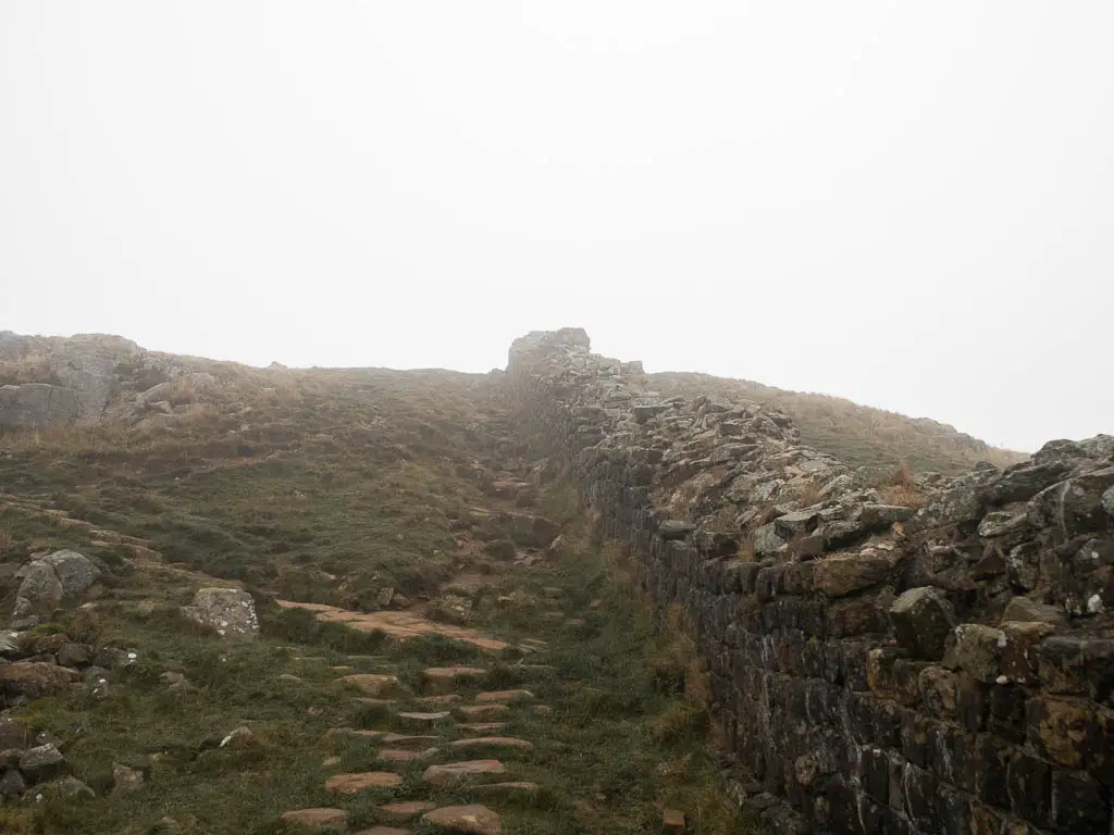 A stone trail running uphill with the stone Hadrian's Wall to the right of it, on a foggy day.