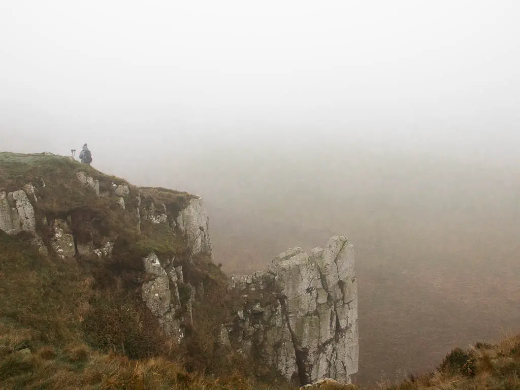 Looking down the side of a rock sticking out from the cliff along the Sycamore Gap walk. There is a man standing on top of the cliff and its a foggy day.