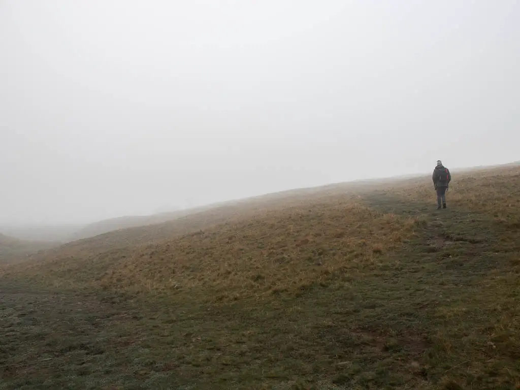 A large grass field, with a man walking on the grass trail leading to the right, on a foggy day.