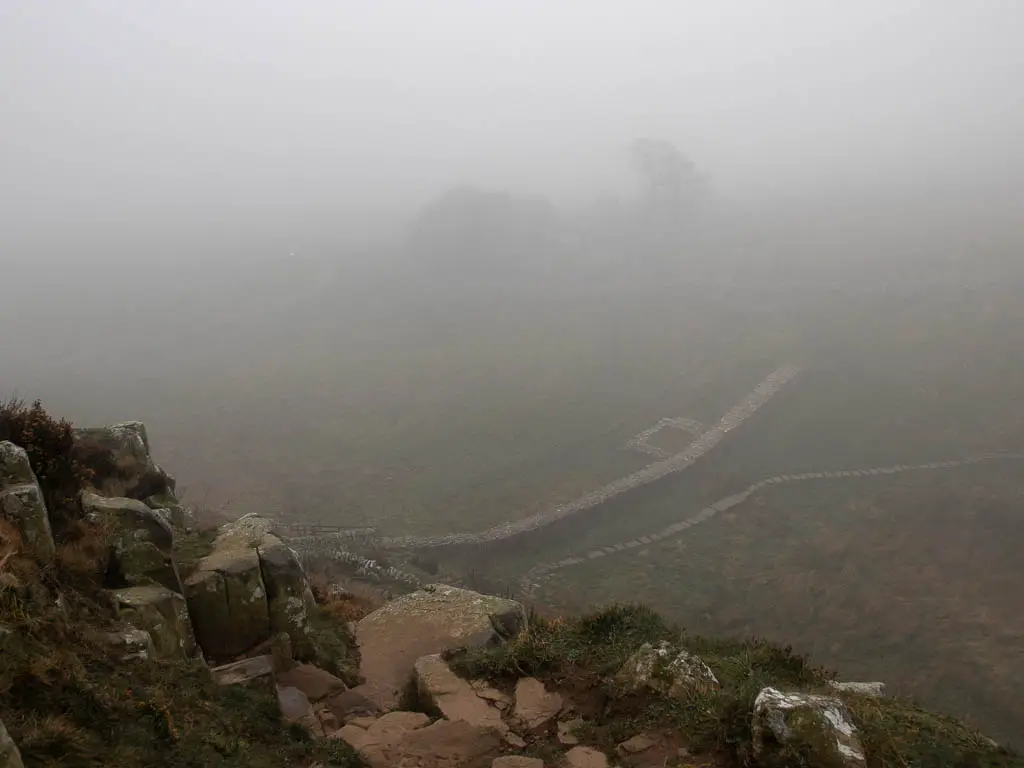 Looking down the rocks of a steep hill,. with Hadrian's wall visible below through the fog.