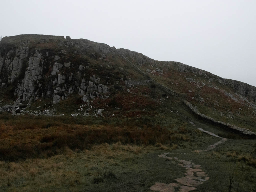 A paved stone trail leading towards a hill, with the stone Hadrian's wall running up it, on the walk towards Sycamore Gap.