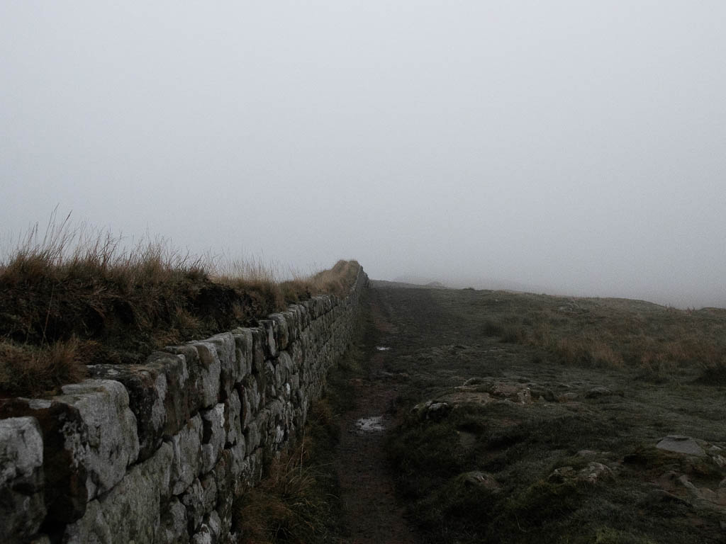 A dirt trail running alongside Hadrian's Wall, on the walk towards Sycamore Gap. The trail and wall are leading into the white fog.