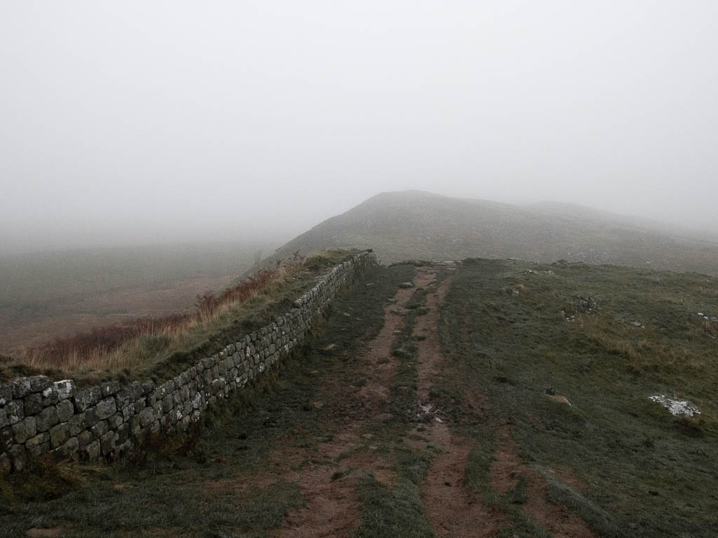 A dirt trail next to Hadrian's Wall, running over a small hill with another hill visible ahead.