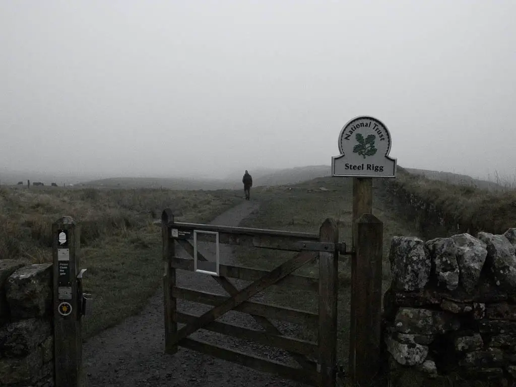 A slightly ajar wooden gate with a national trust sigh saying 'Steel Rigg' at the start of the walk to Sycamore Gap. There is a man walking ahead on the trail. the air is dark and foggy.