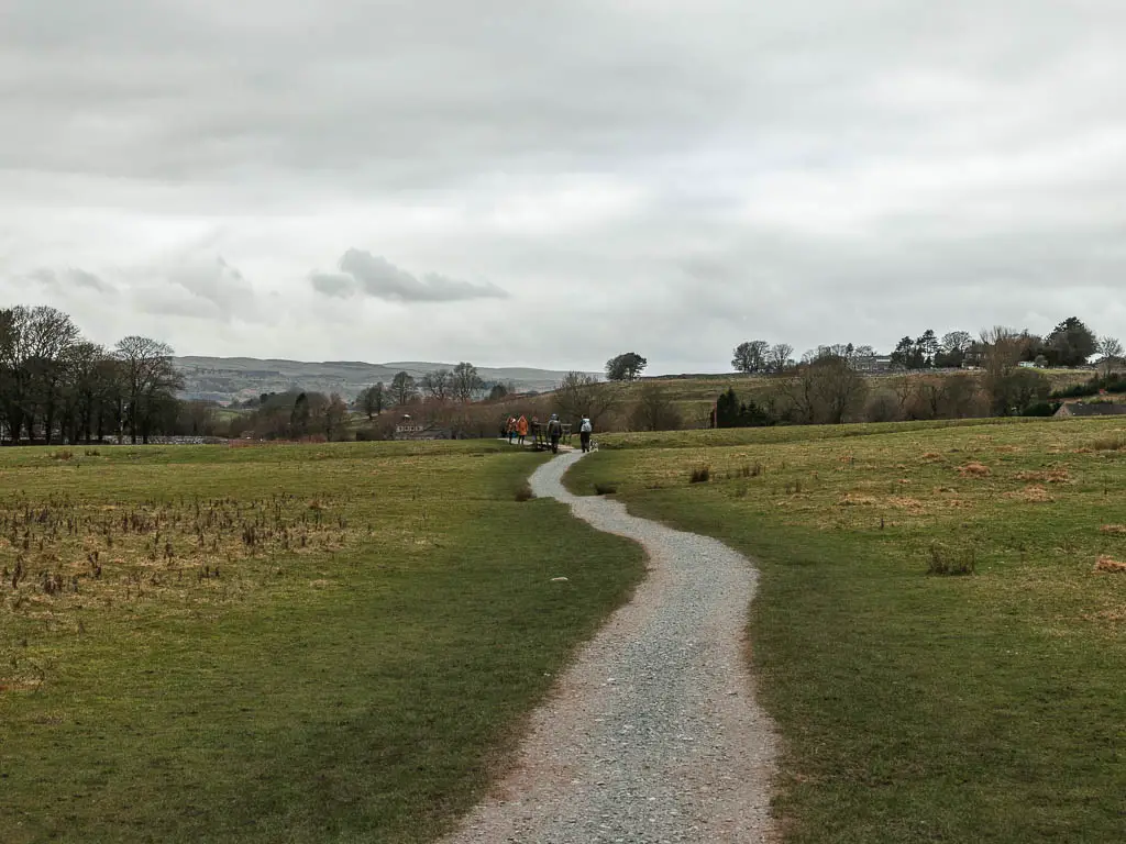 A gravel path snaking through a large grass field, with a group of people walking on it ahead. 