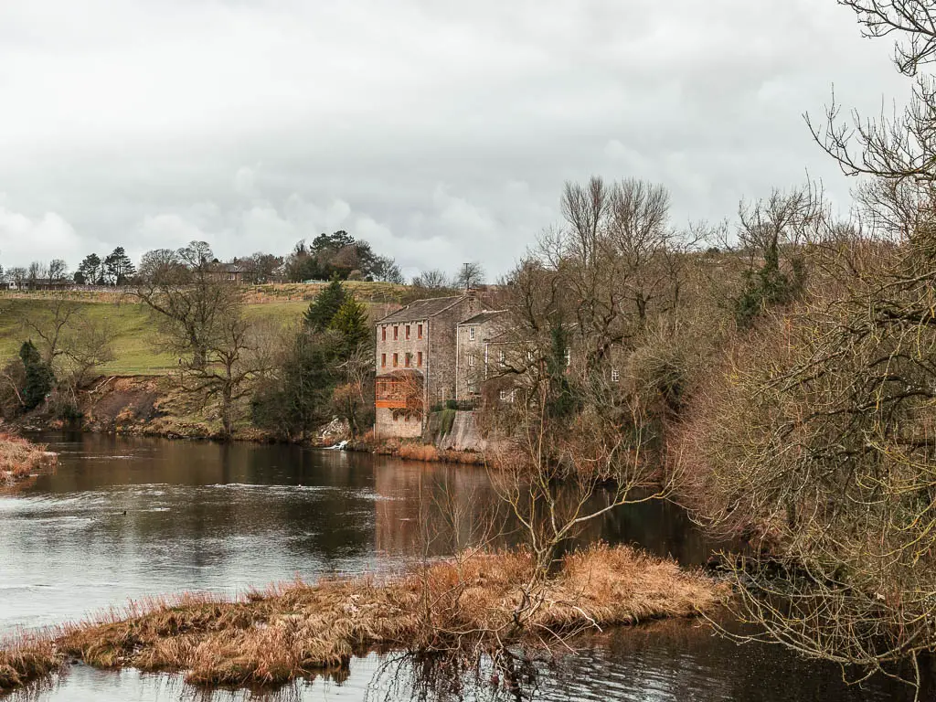 Looking across the river to a house perched on the edge, nestled within the leafless trees, on the walk from Burnsall to Grassington.