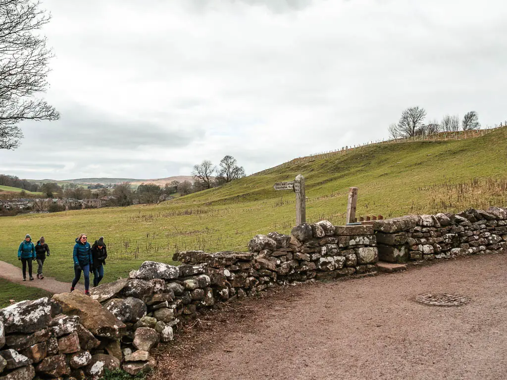 A stone wall along the left side of the road, with a wooden trail signpost pointing into the grass field on the other side. There are people walking this way towards the stone wall.