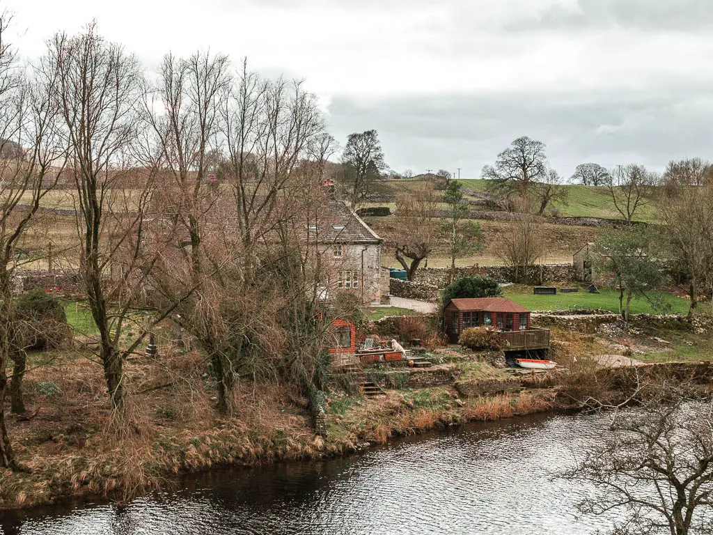 Looking across the river towards a stain house partially hidden by leafless trees on the walk towards Grasisngton from Burnsall.