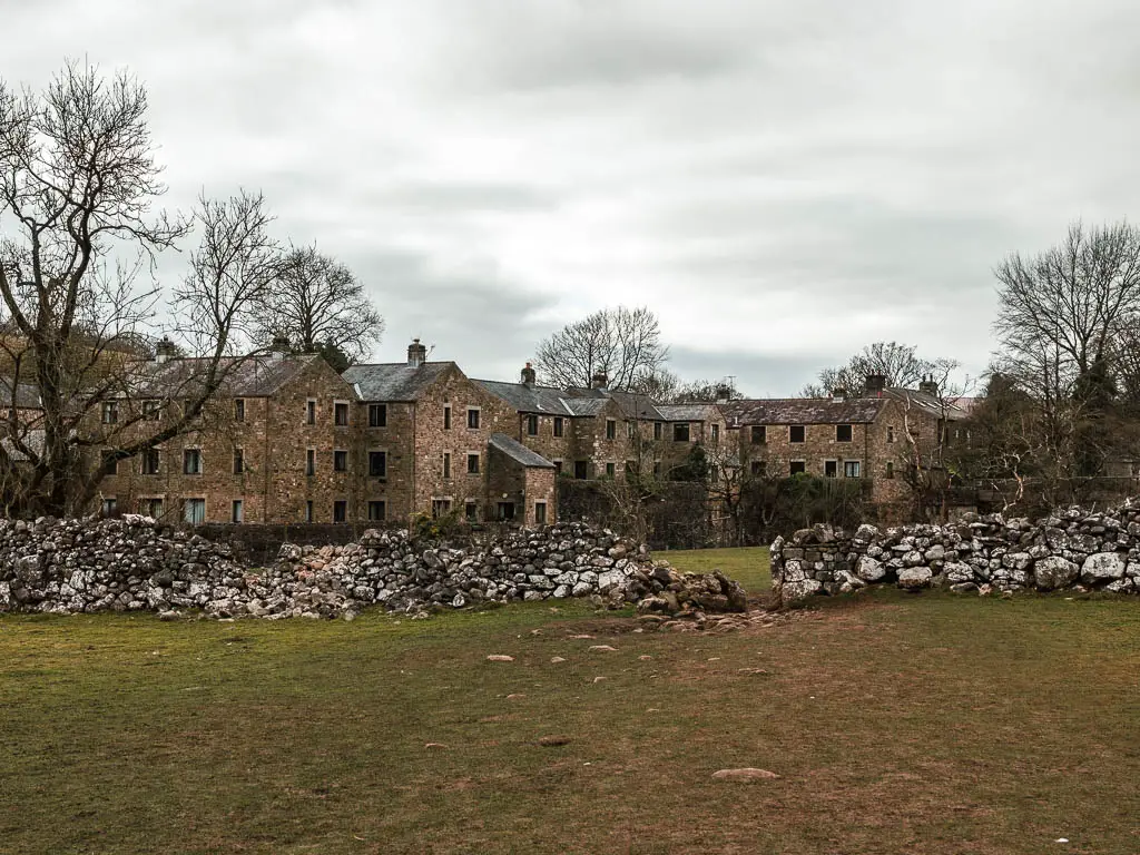 A stone wall with a gap on the other side of the grass field with a mass of stone buildings ahead on the other side.