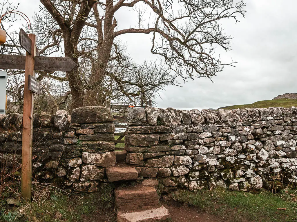 Stoner steps leading up through a stone wall. There is a wooden trail signpost on the left side.