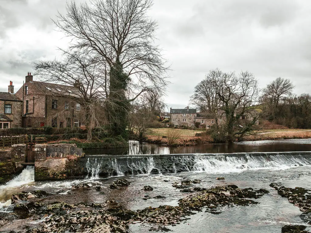 Looking across the river to the Linton Falls, on the walk into Grassington from Burnsall. There are brown coloured stone houses on the other side and a few big leafless trees.