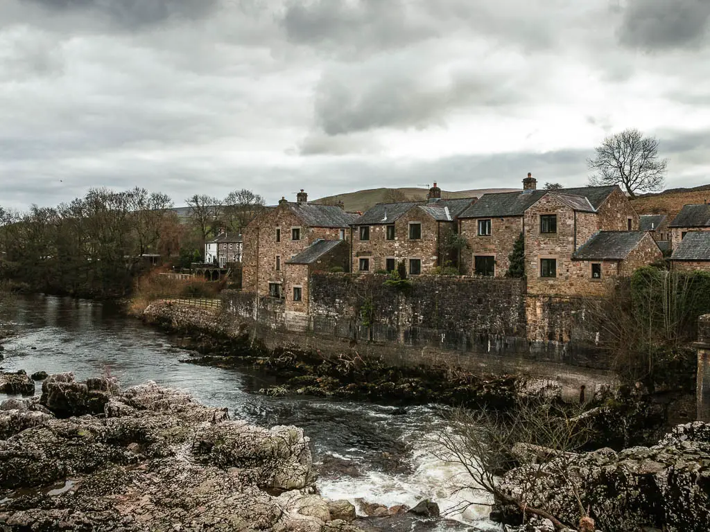 Looking across the river and falls, surround by rocks, and the village of Linton on the other side half way through the Burnsall to Grassington walk. The sky is grey.