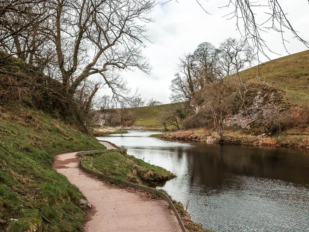 A well maintained neat path snaking along the left side of the river on the walk out from Burnsall towards grassington. There is a green grass hill on the other side of the river. 