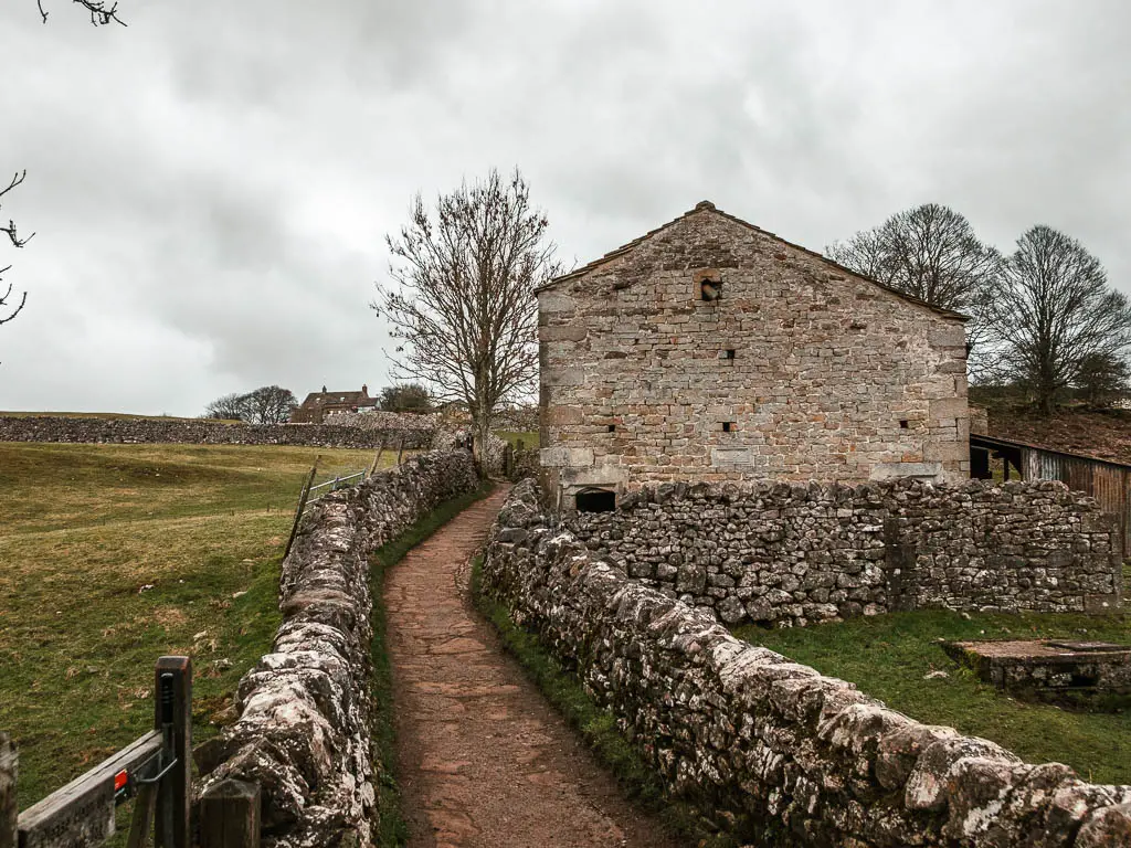 A narrow stone path lined with a stone wall on the walk up to Grassington from the Linton Falls. There is a stone walled building on the right side of the wall.