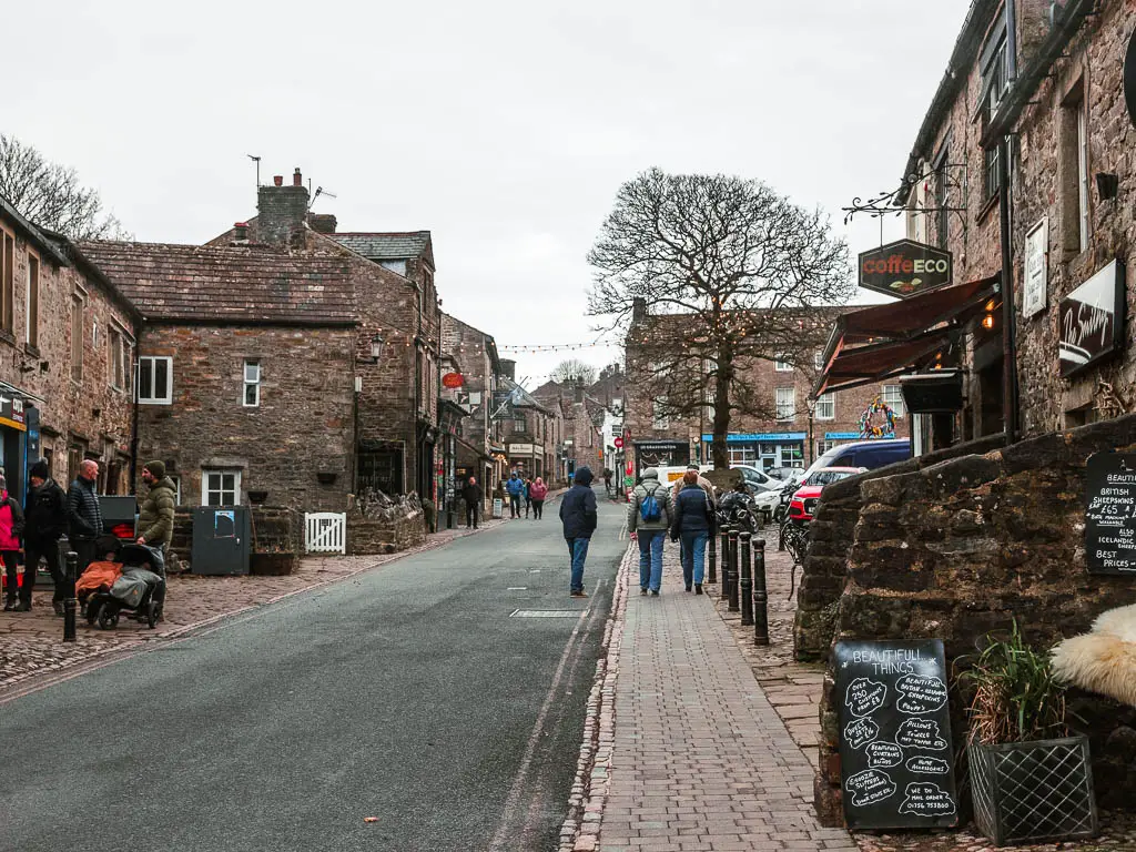 Looking along Main Street in Grassington, lined with stone building shops. There are lots of people walking along it.