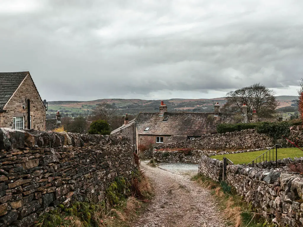 Looking down along the gravel path, towards stone houses, and hills visible in the distance.  There is a tall stone wall on the left and a short one on the right. 