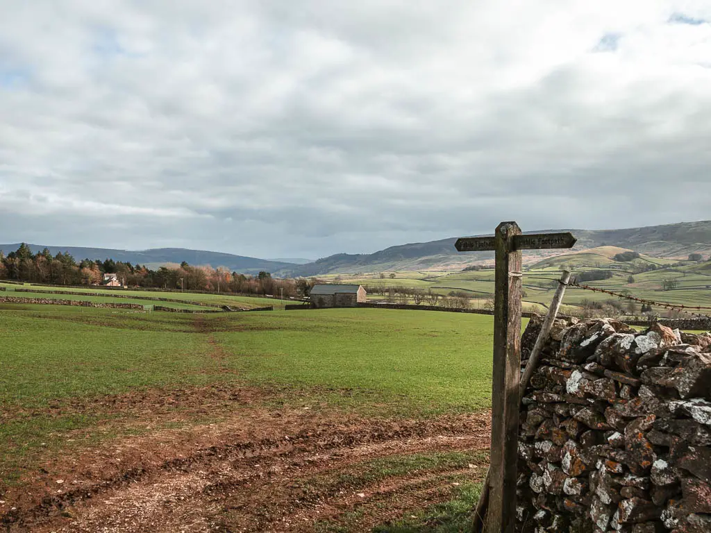 A wooden trail signpost on th right, pointing ahead across a large green grass field. There is a shed ahead in the distance in the field.